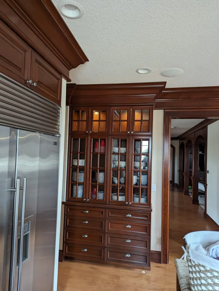 A built-in wooden cabinet with glass doors and multiple drawers is set against a wall next to a stainless steel refrigerator in a home interior.