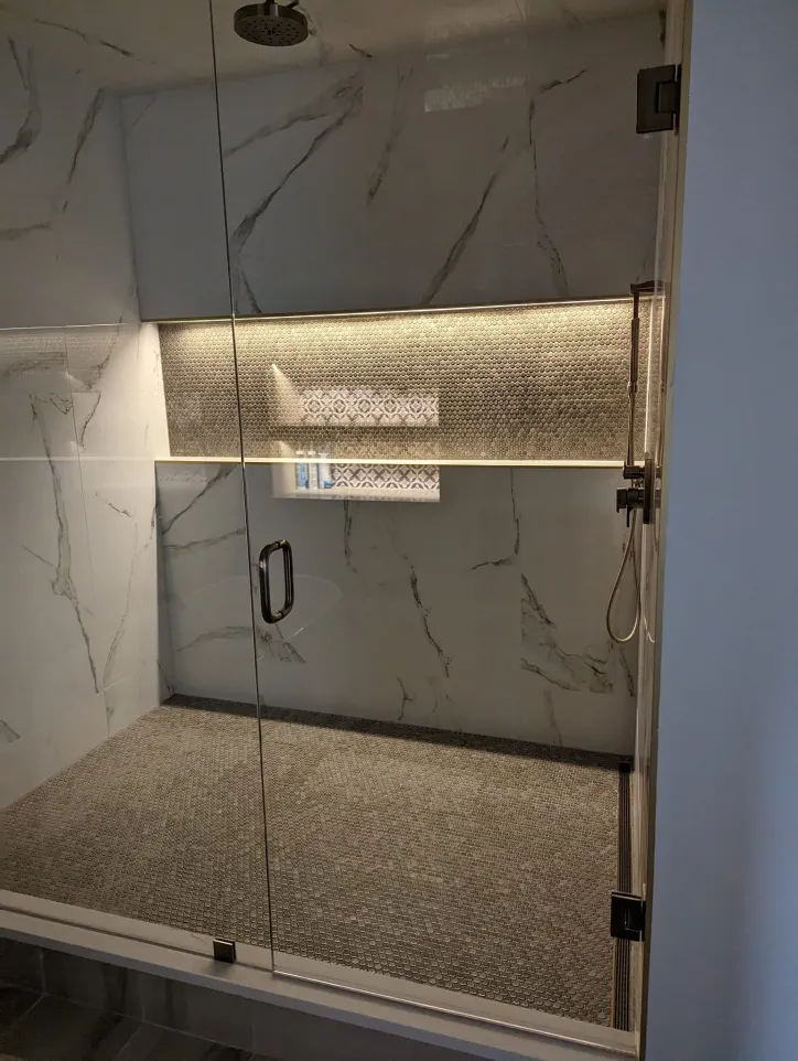 Modern glass shower with marble walls, tiled floor, built-in shelf with mosaic backsplash, and an overhead light above the shelf, viewed from outside the enclosure.