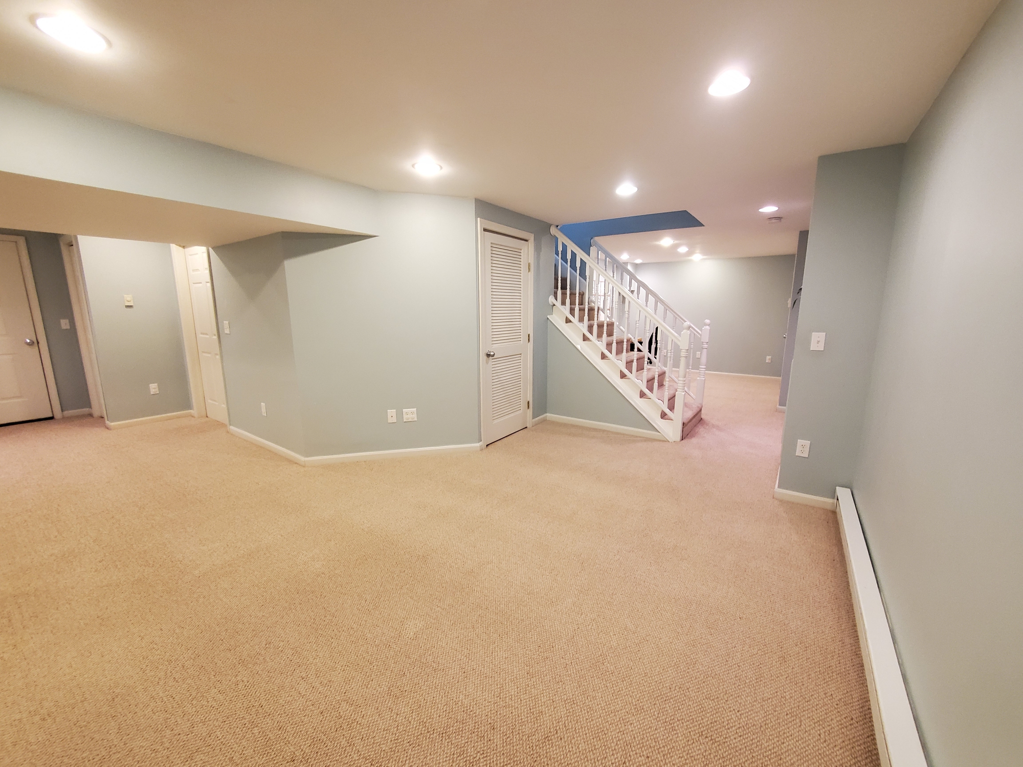 Spacious finished basement with beige carpet, light blue walls, white trim, and a staircase leading upstairs; multiple doors and recessed ceiling lights are visible.