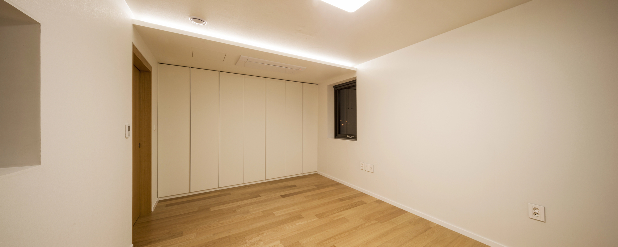 Empty room with light wood flooring, white walls, a closed closet, a small window, and ceiling light. Electrical outlets are visible near the floor.