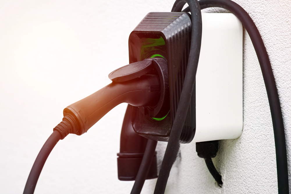 Close-up of an electric vehicle charging plug connected to a wall-mounted power outlet, with cables looped around the charger.