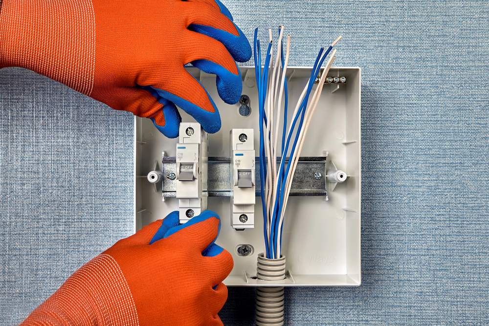 Hands in orange gloves installing circuit breakers and connecting electrical wires inside a white breaker box mounted on a textured blue wall.