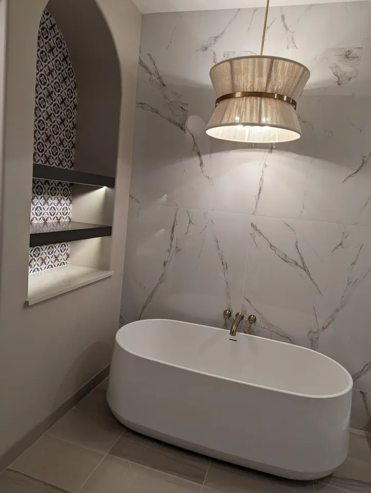 Modern bathroom with a white freestanding tub, marble wall tiles, a large pendant light, and a niche with patterned tiles and shelves.